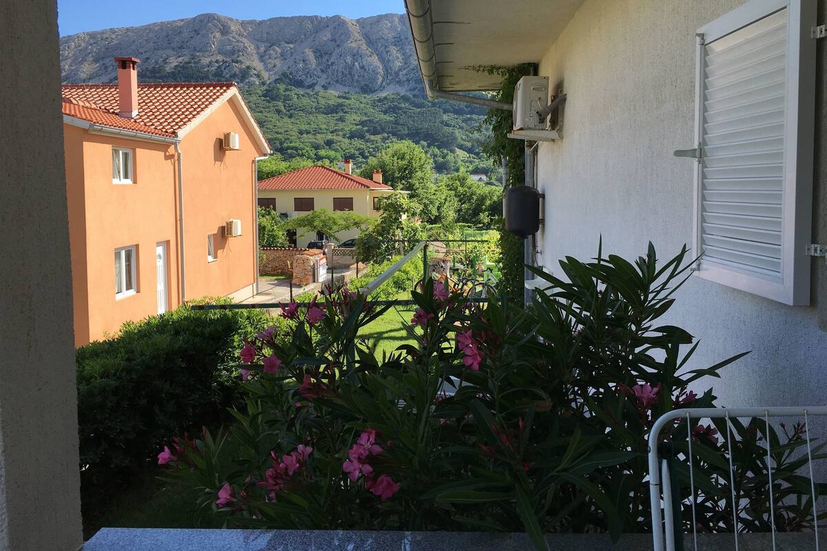 A balcony with a view of a mountain, a building with a red roof, and a garden with pink flowers.