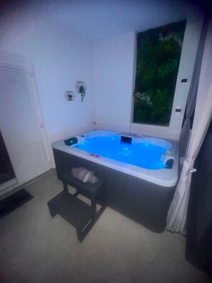 A hot tub with a step, a window with a view of greenery, and a white wall.