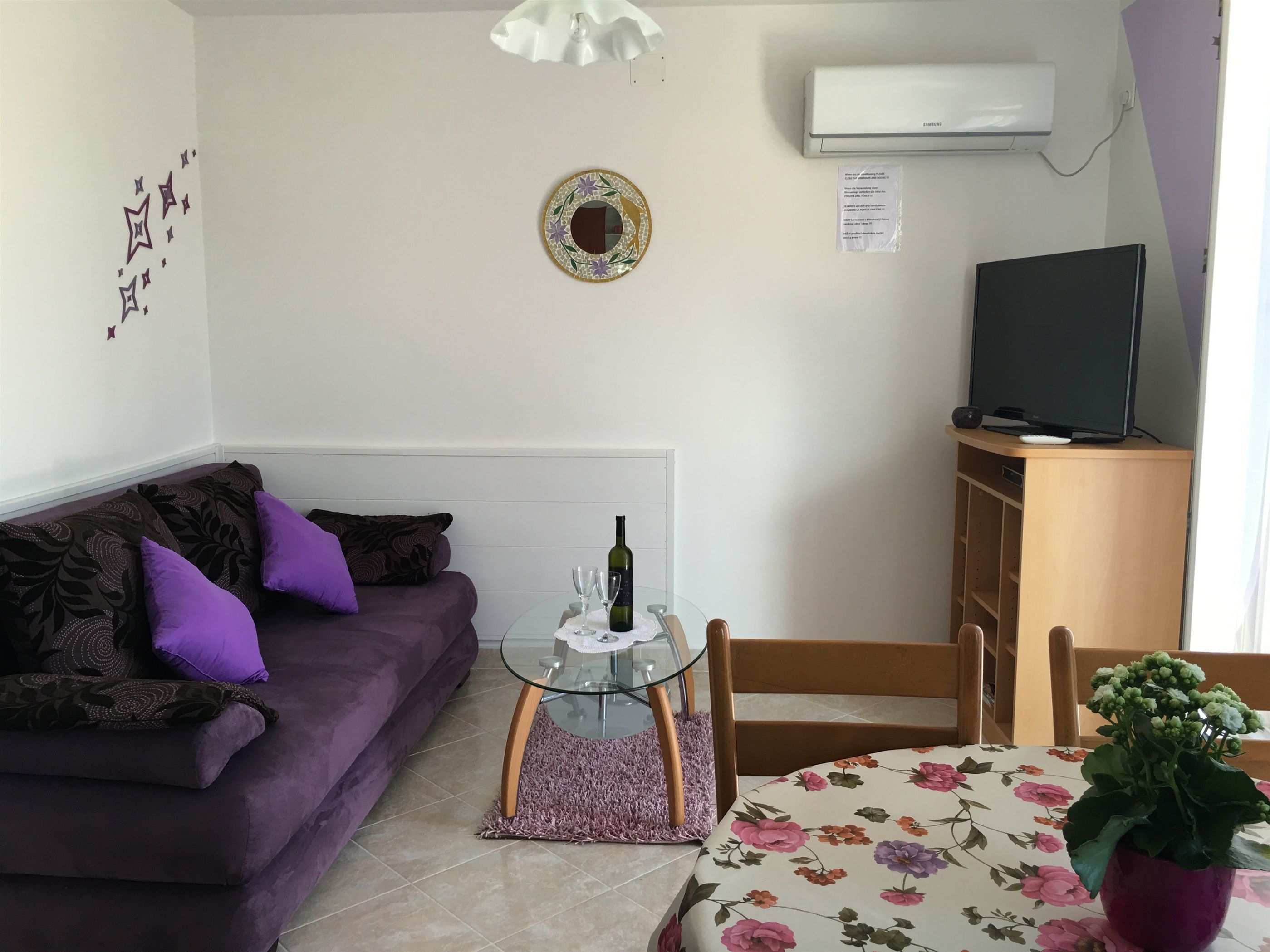 Een woonkamer met een paarse bank, een glazen bijzettafeltje, een tafelkleed met bloemenprint, een kamerplant, een televisie op een houten standaard, en een airconditioner aan de muur.