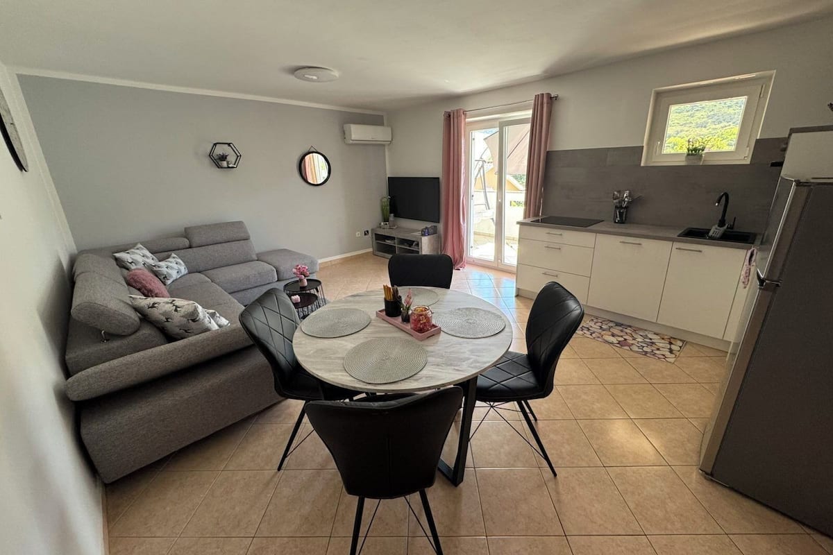 A compact living space with a grey sofa, a dining table with chairs, a kitchen area with a sink and a window, and a flat-screen TV mounted on the wall.