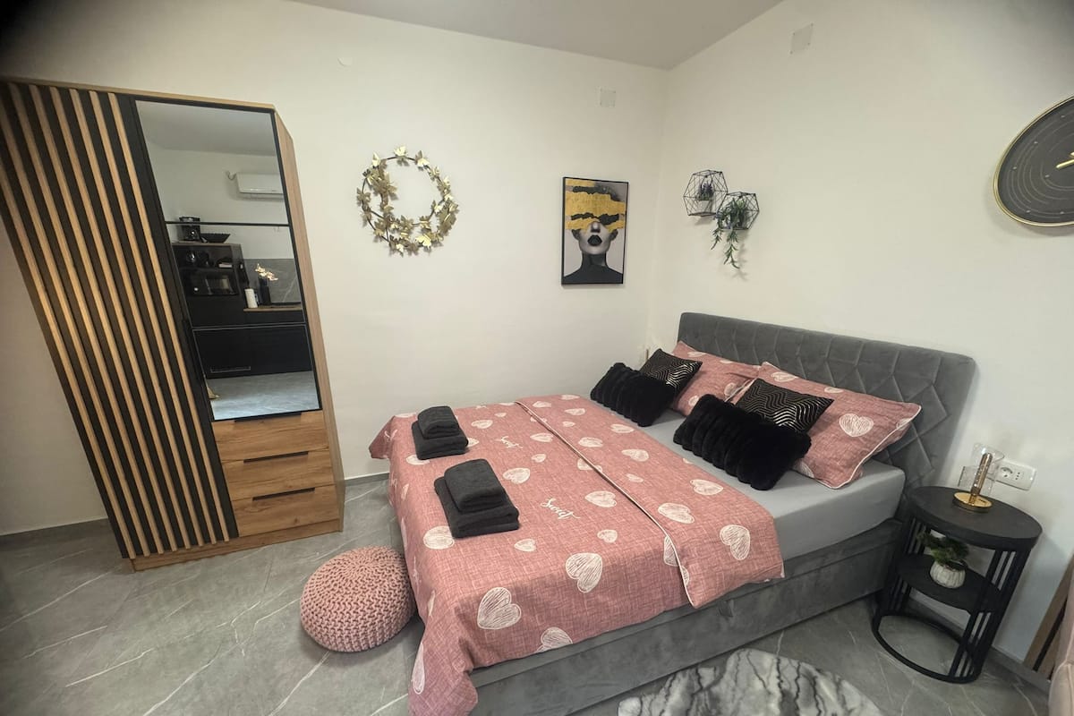 A bedroom with a bed, a wardrobe, a mirror, a wall art piece, and a small side table with a plant.