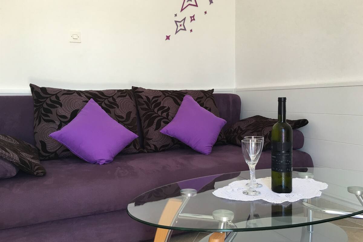 A purple sofa with patterned cushions, a glass table with a wine bottle and glass, and a wall with star decals.