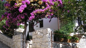 Un mur de pierre surmonté d’une rambarde métallique, un arbre aux fleurs violettes et un bâtiment doté d’un toit en pente et de volets.