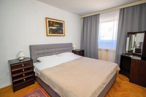 Ein Schlafzimmer mit Bett, Nachttischen, einer Lampe, einer Kommode, einem Teppich, einem Bild und einem Fenster mit Vorhängen.