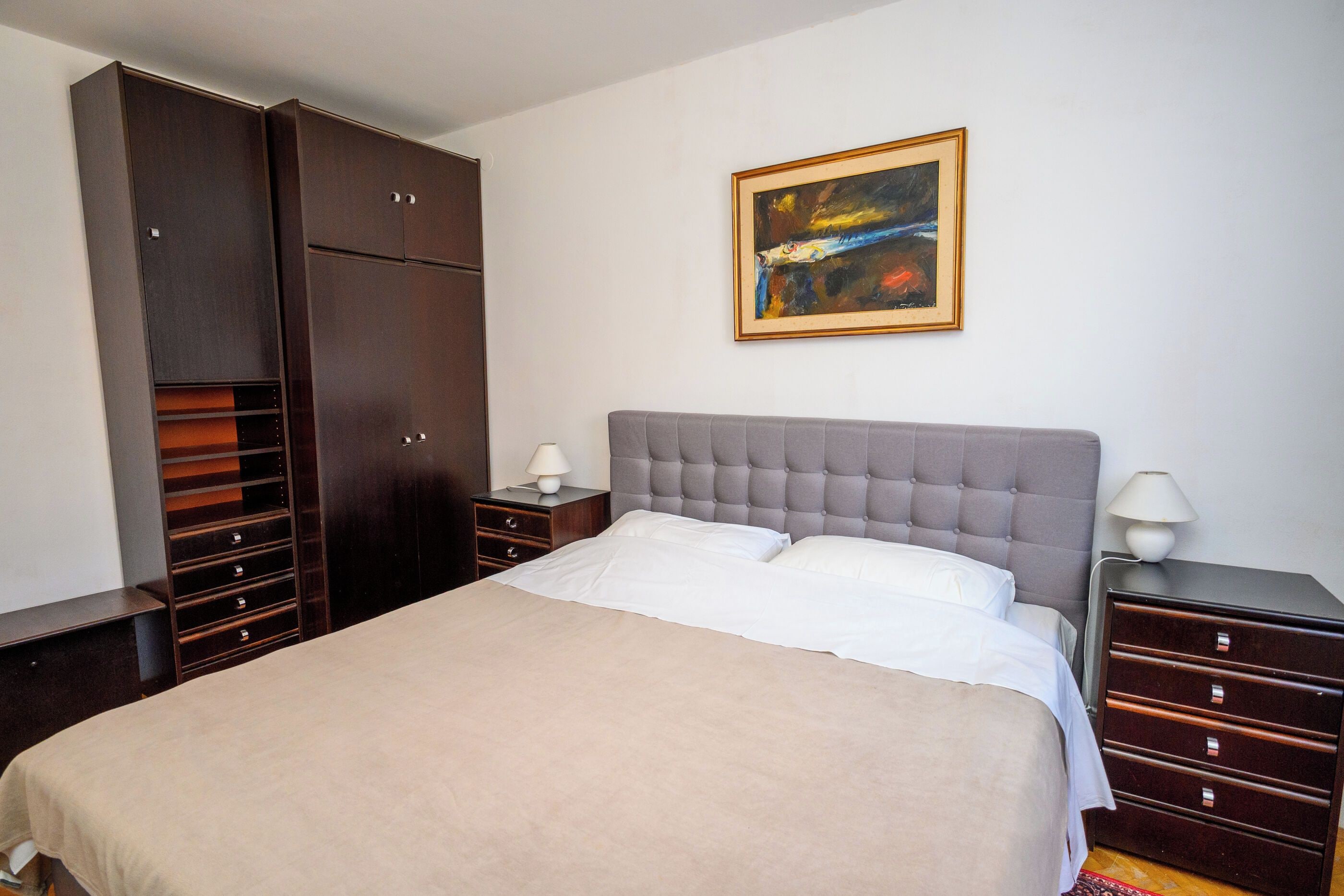 Un dormitorio con una cama grande, un armario, una mesita de noche, una lámpara y un cuadro en la pared.
