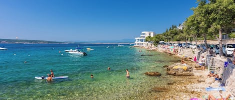 Una playa con agua cristalina, gente nadando y tomando el sol, y un edificio con balcón con vista al mar.
