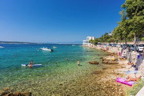 A beach with clear water, people swimming and sunbathing, and a building with a balcony overlooking the sea. - Apartment Kralj (Selce)