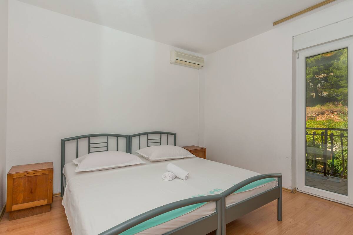 Ein Schlafzimmer mit Bett, zwei Nachttischen, einem Fenster mit Blick ins Grüne und einer Klimaanlage.