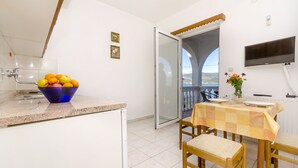 A kitchen with a bowl of fruit on the counter, a dining table with chairs, a TV mounted on the wall, and a balcony with a view. - Apartments Tomislav (Slatine)