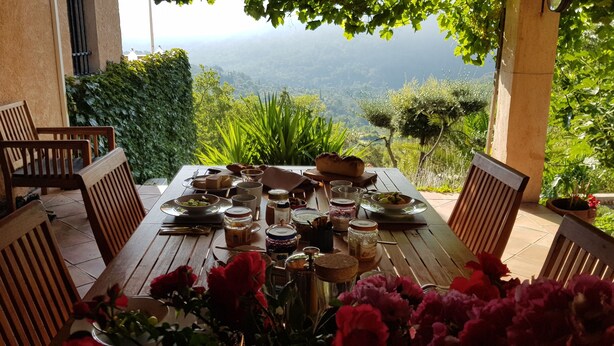 A wooden table set for a meal with various dishes and utensils, surrounded by chairs, with a view of greenery and hills in the background. - L'Estuve Provençale (Bargemon)