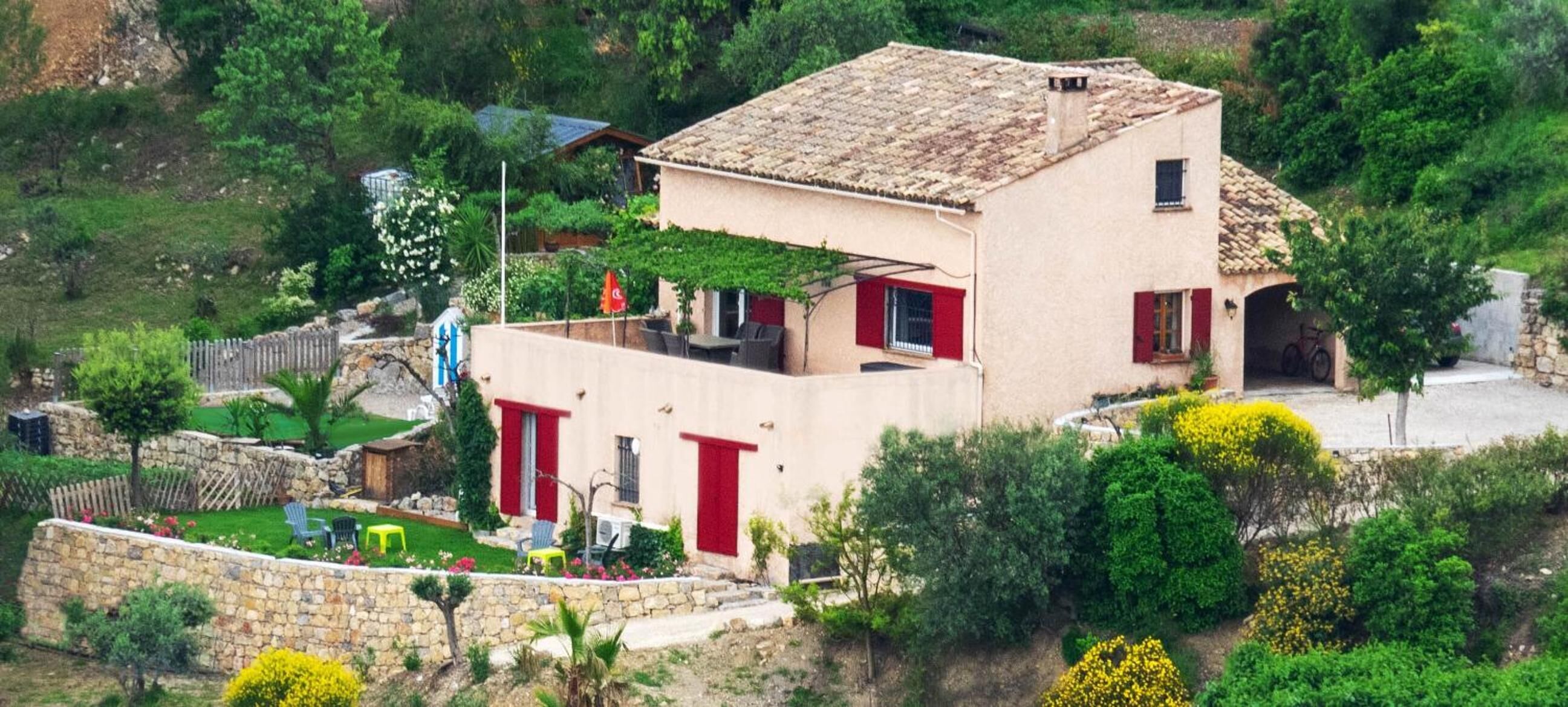 Une maison à deux étages, dotée d’un toit plat, de volets rouges et d’un petit balcon.