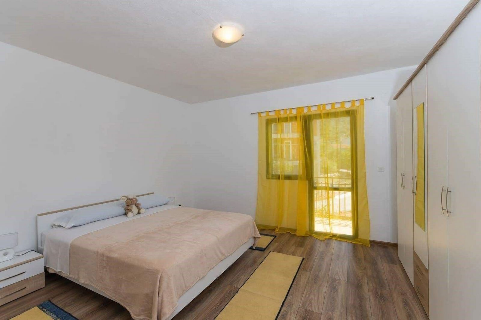 A bedroom with a bed, a nightstand, a wardrobe, and a window with yellow curtains.