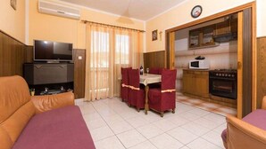 Ein kompakter Wohnbereich mit Sofa, Esstisch mit vier Stühlen, Fernseher, Küche mit Mikrowelle und Backofen sowie einer Wanduhr.