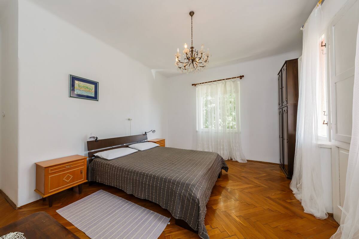 Un dormitorio con suelo de madera, una cama con colcha estampada oscura, una mesita de noche, un aparador, una araña y una ventana con cortinas de encaje.