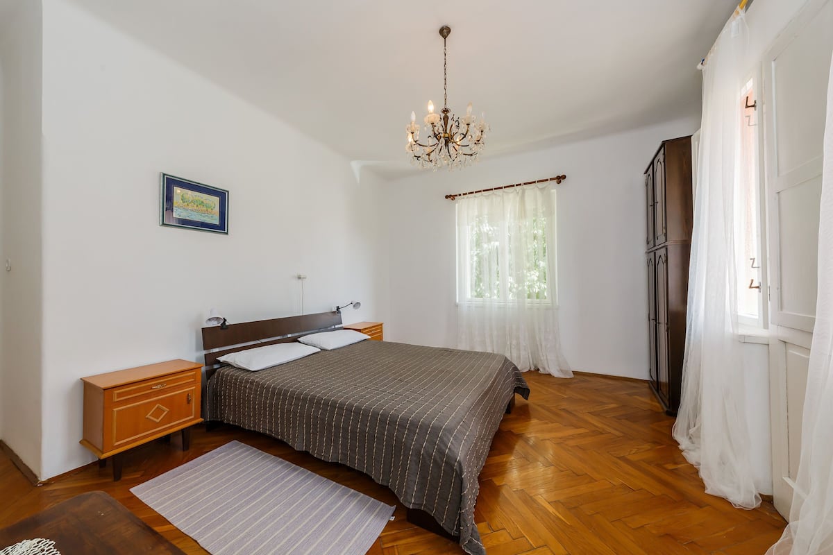 Un dormitorio con suelo de madera, una cama con colcha estampada oscura, una mesita de noche, un aparador, una araña y una ventana con cortinas de encaje.