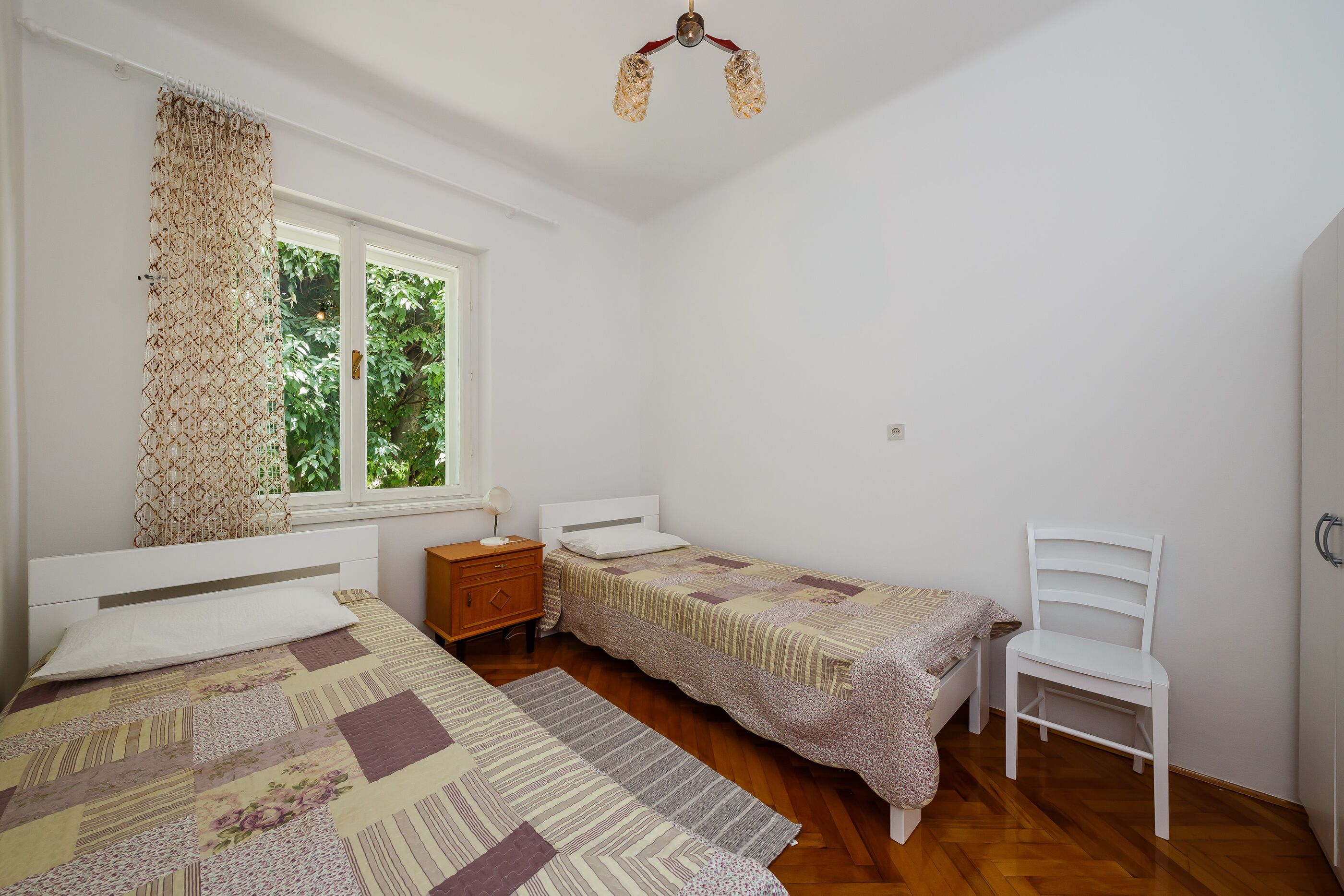 A bedroom with two single beds, a wooden dresser, a white chair, and a window with a view of greenery.