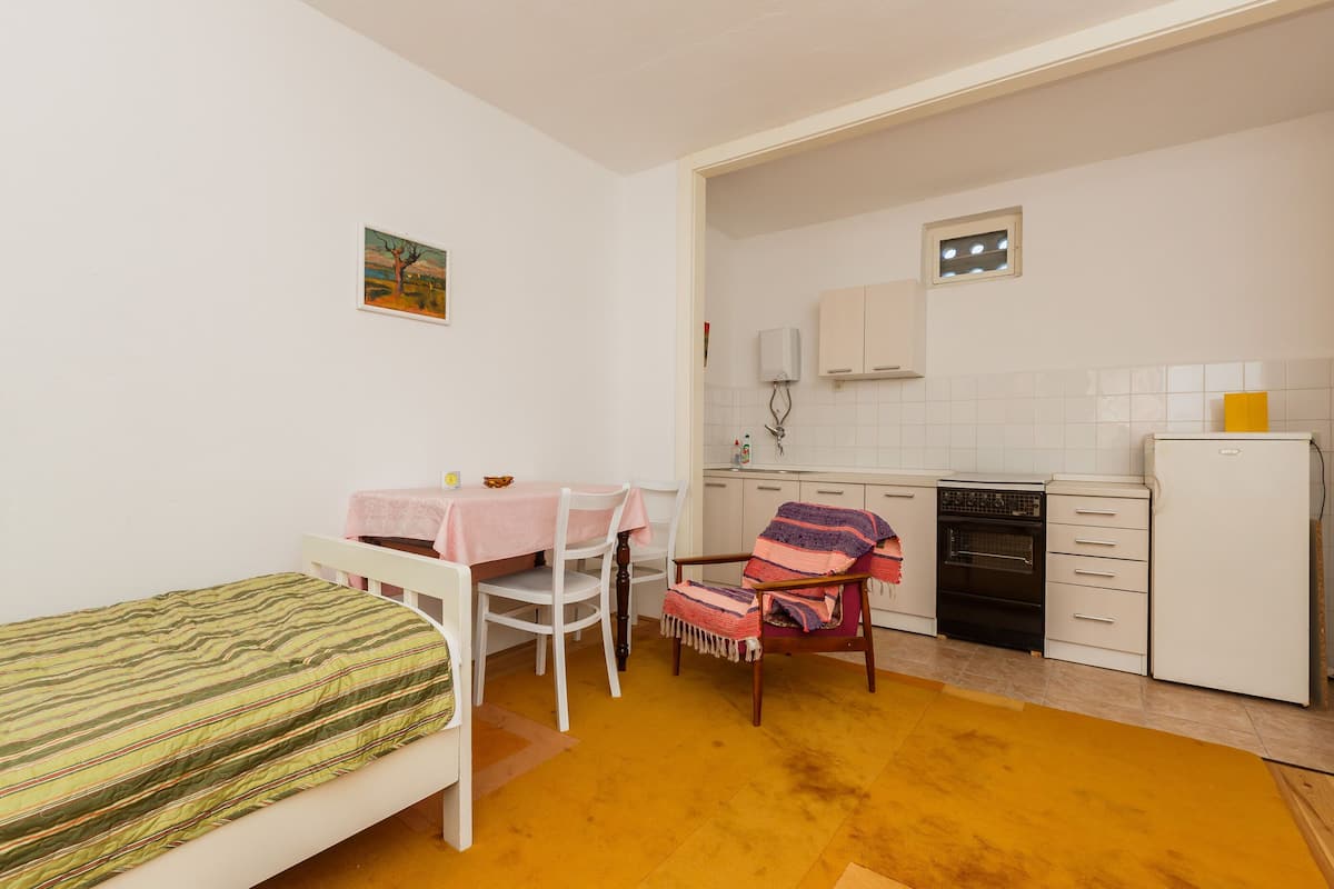 Una habitación pequeña y funcional con cama, una mesa de comedor con sillas, y un área de cocina con refrigerador, horno y armarios.