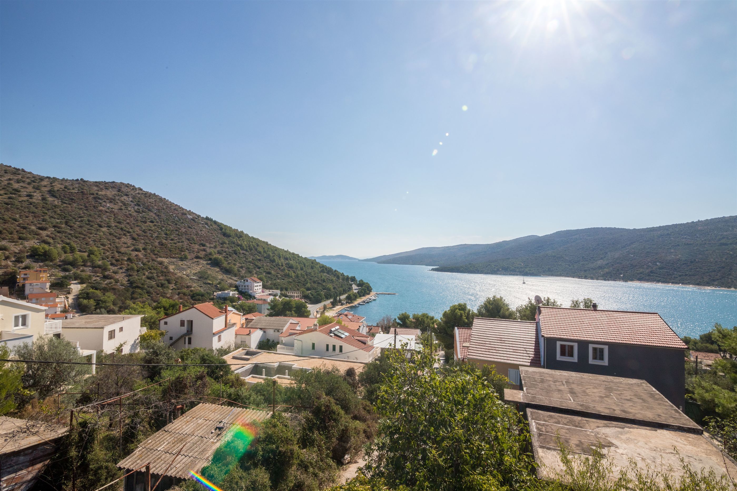 A coastal village with houses, a clear blue sea, and hills in the background.