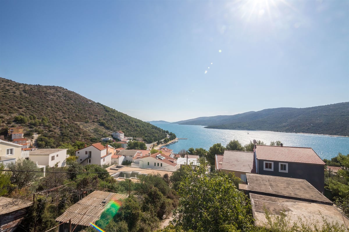 A coastal village with houses, a clear blue sea, and hills in the background.
