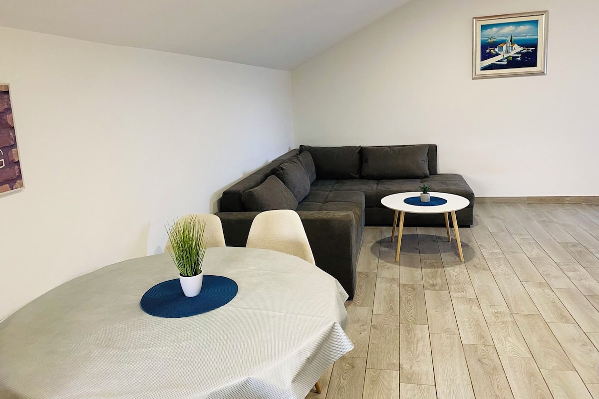 A modern living room with a sectional sofa, a round dining table with a plant, and a small side table.