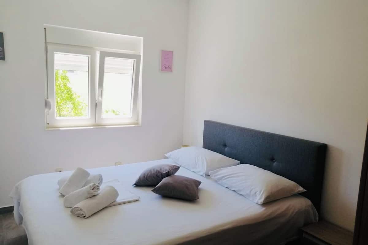A neatly made bed with white linens and rolled towels, a grey upholstered headboard, and a window with blinds.