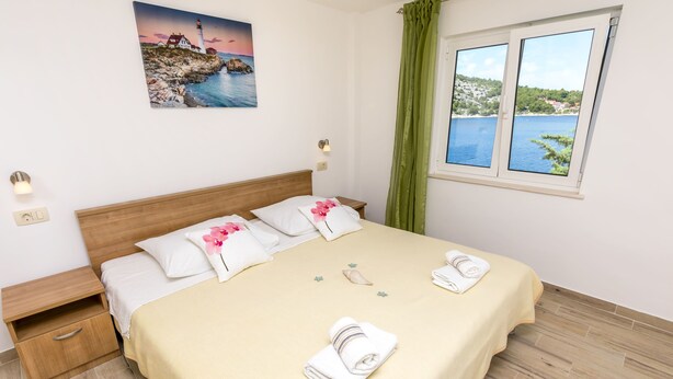 A neatly made bed with a beige bedspread, two pillows, and two folded towels. There is a nightstand with a drawer, a wall-mounted light, and a painting of a coastal scene. A window with a view of a body of water and greenery is visible. - Apartment Vukorepa (Razanj)