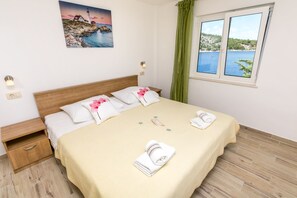 A neatly made bed with a beige bedspread, two pillows, and two folded towels. There is a nightstand with a drawer, a wall-mounted light, and a painting of a coastal scene. A window with a view of a body of water and greenery is visible. - Apartment Vukorepa (Razanj)