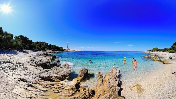 A beach with clear water, rocky shore, and a lighthouse in the distance. - Apartment Ivan (Veli Rat)