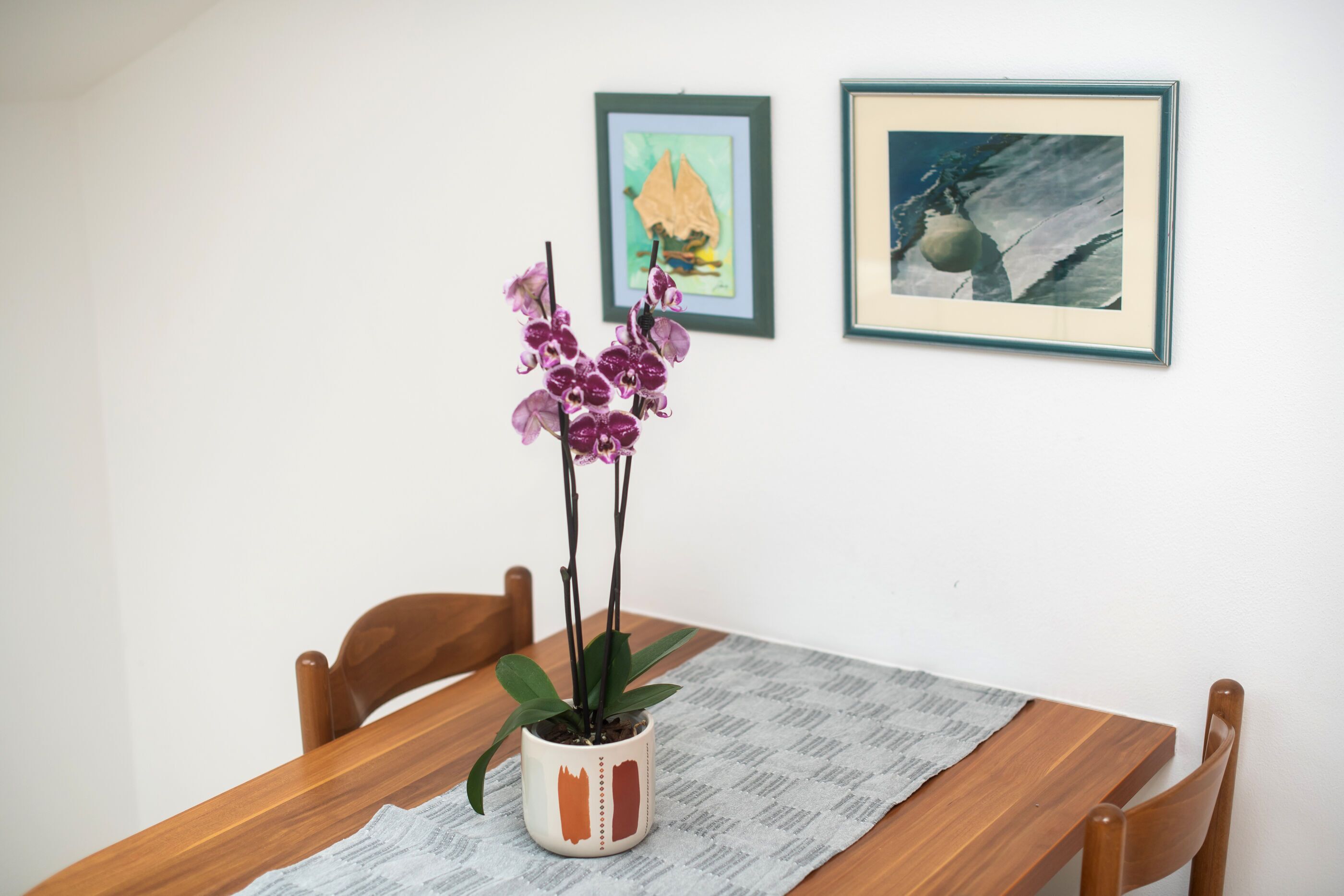 A wooden dining table with a potted orchid and two framed artworks on the wall.