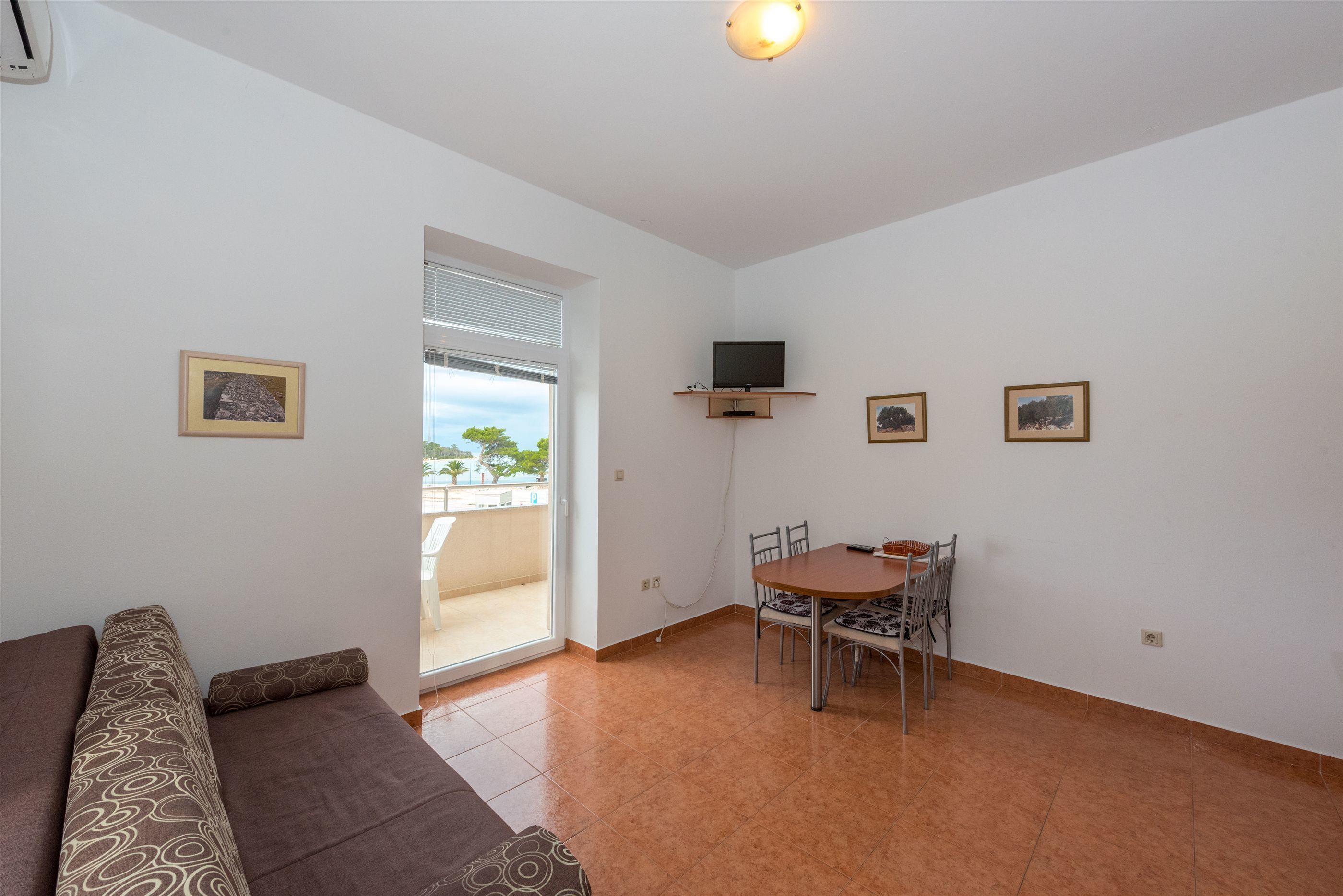 A living room with a sofa, a dining table with chairs, a TV mounted on the wall, and a door leading to a balcony.
