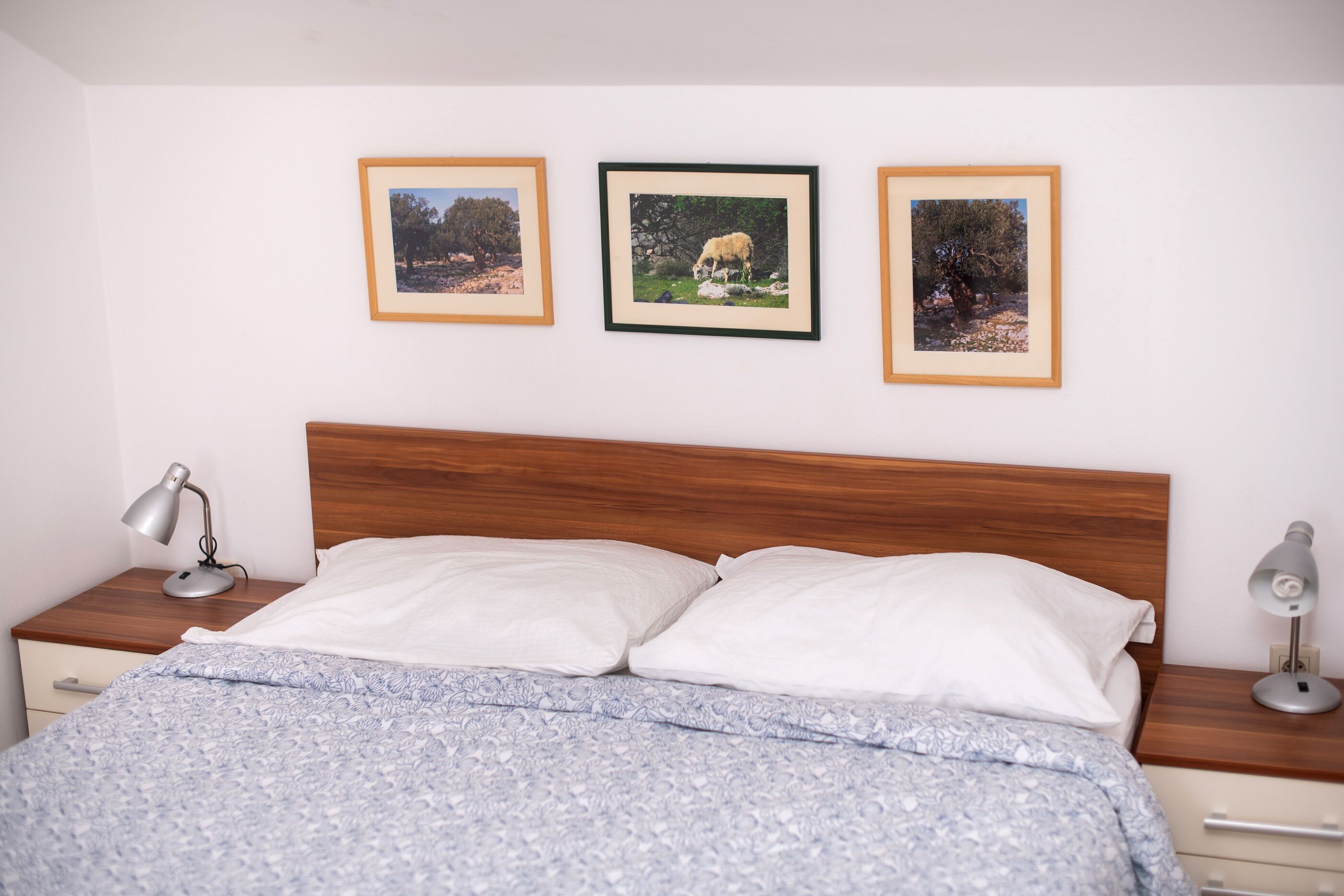 A bedroom with a wooden headboard, a bed with white and patterned bedding, and three framed pictures on the wall.