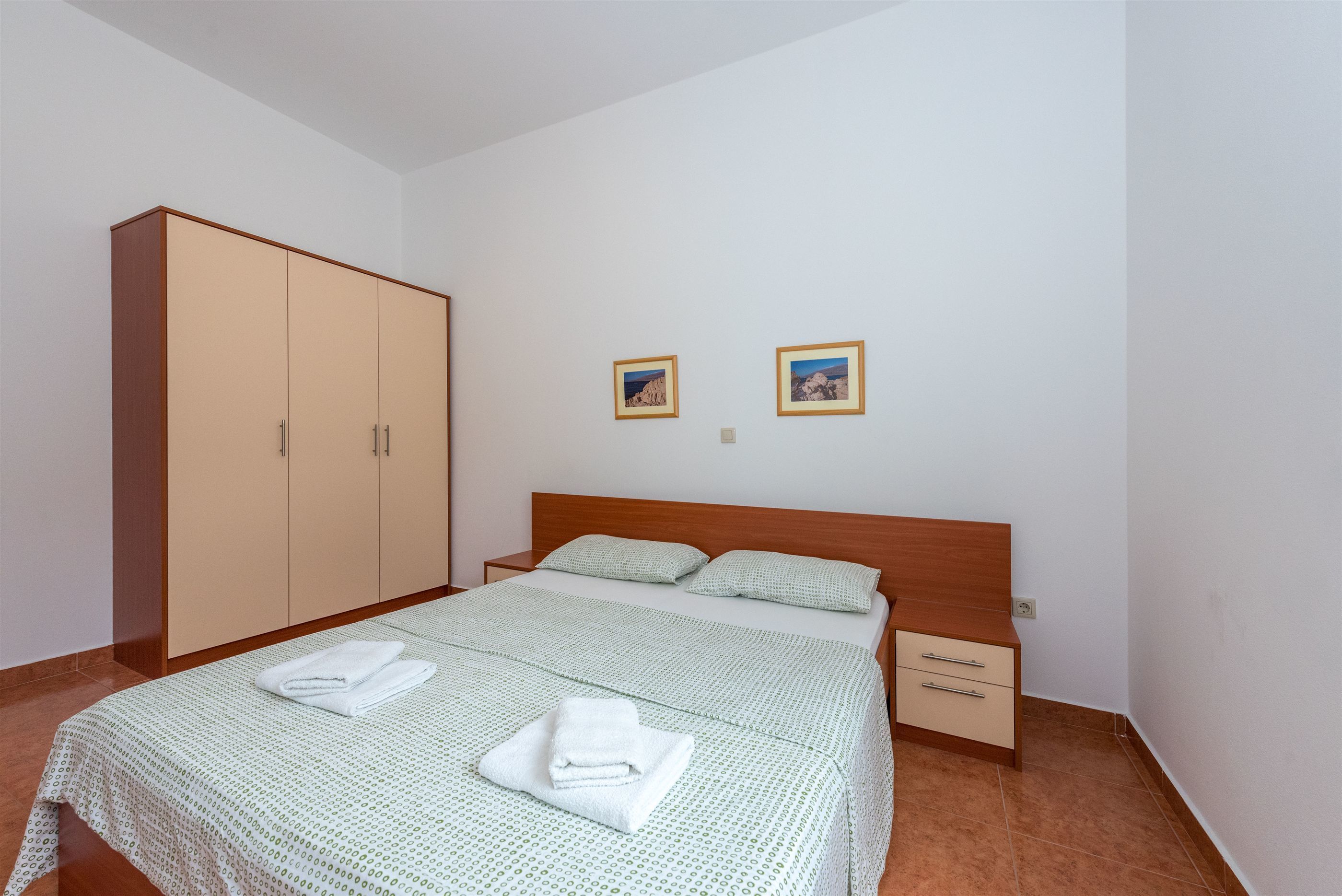 A bedroom with a bed, a wardrobe, two framed pictures on the wall, and a nightstand.