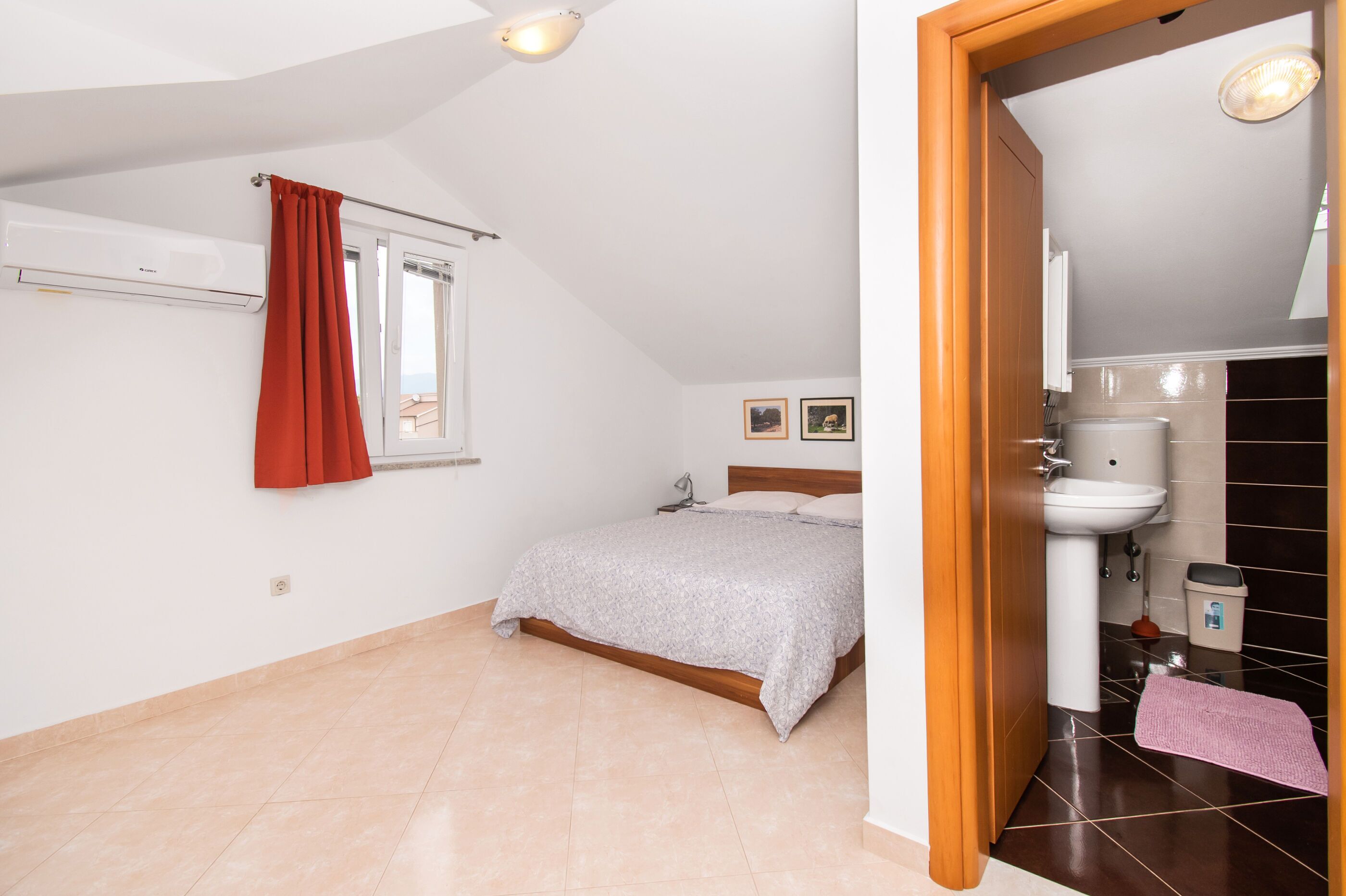 A bedroom with a bed, a window with a red curtain, a white air conditioner, and a view of the outside.