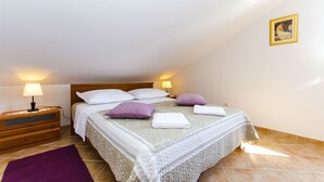 A neatly made bed with white and purple pillows, a wooden nightstand with a lamp, and a framed picture on the wall. - Apartment Lavanda (Rogoznica)