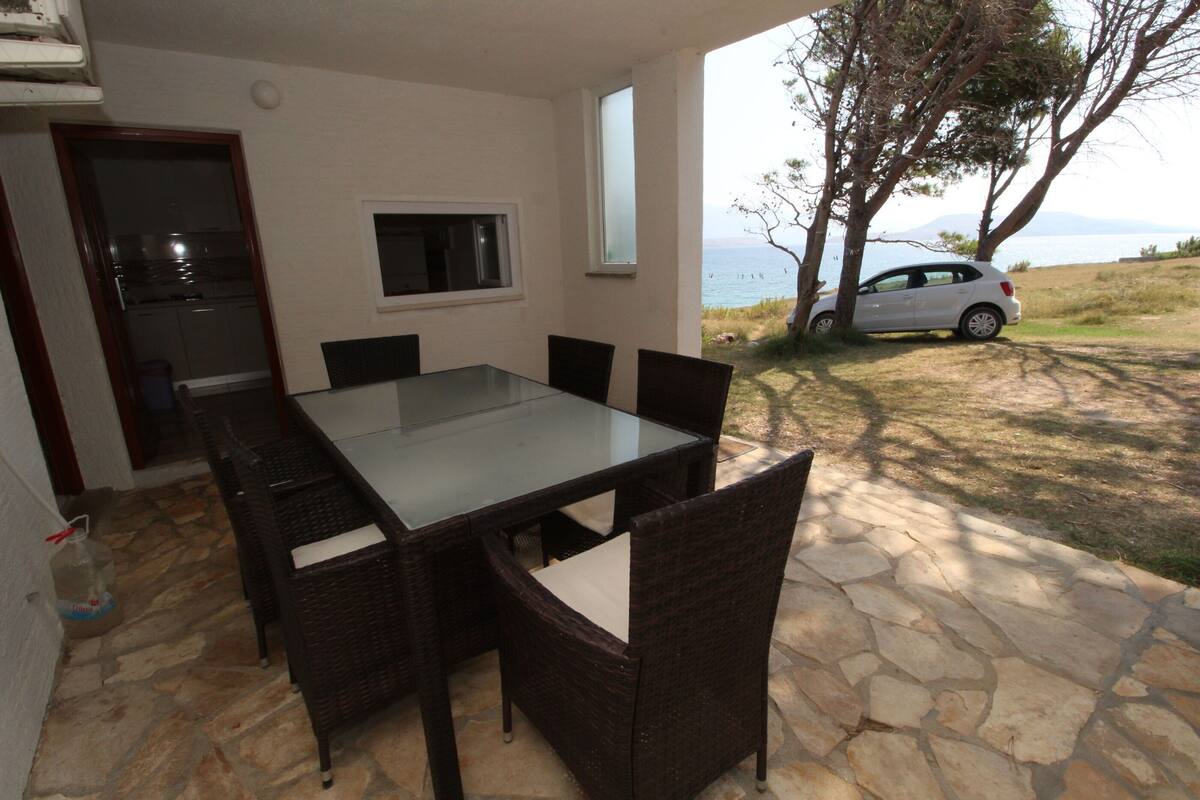 Un coin repas avec une table à plateau en verre et des chaises en osier, une fenêtre et une vue sur une voiture et le paysage.