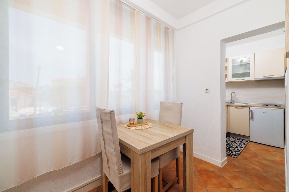 A small dining area with a wooden table and chairs, a window with sheer curtains, and a kitchen with white cabinets and a tiled backsplash.