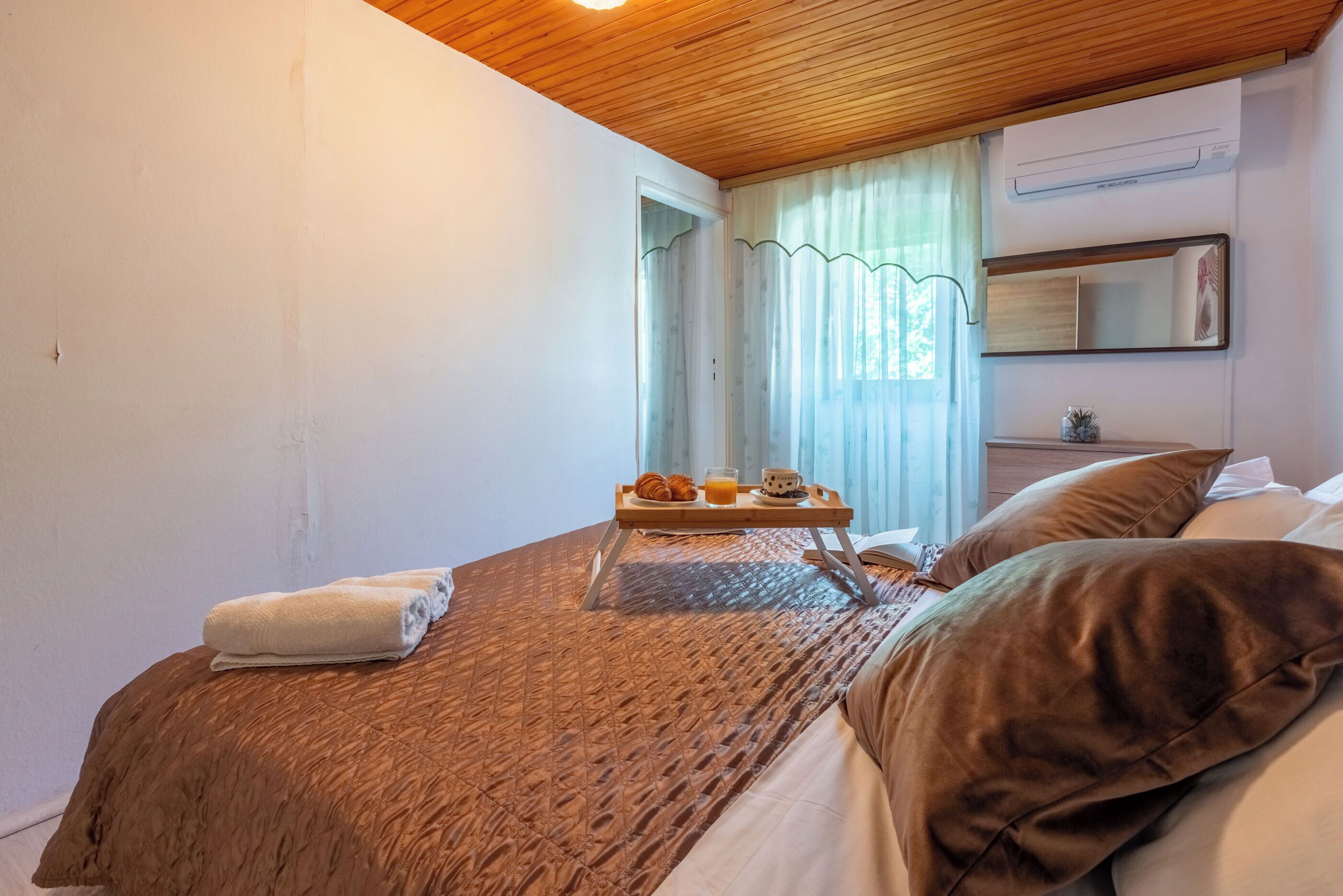 Camera d'albergo con soffitto in legno, un letto con biancheria marrone, un comodino con un vassoio di prodotti per la colazione, una finestra con tende e un climatizzatore a parete.