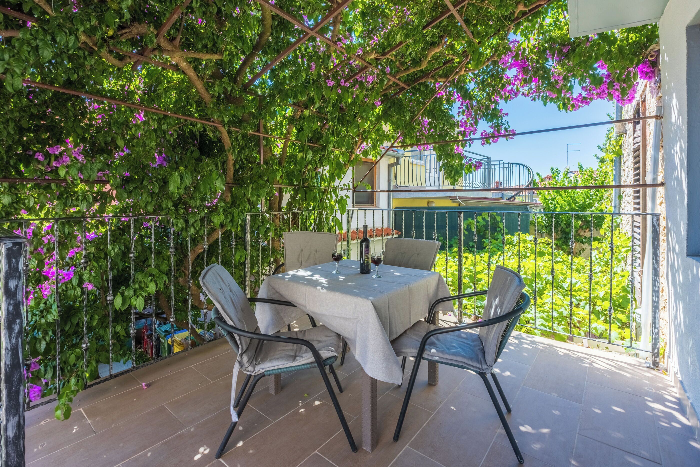 An outdoor patio with a table and chairs, surrounded by greenery and a view of a building.