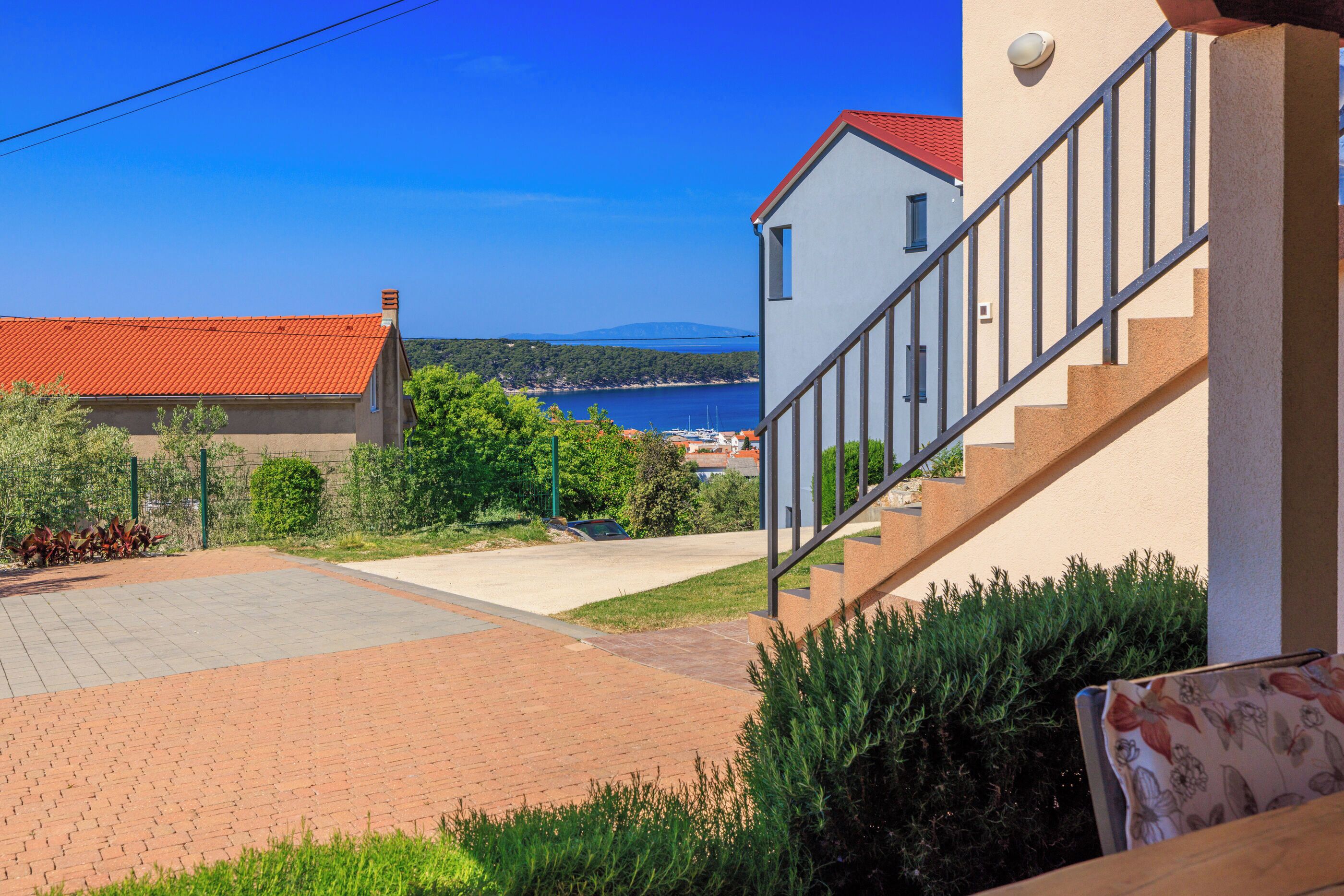 A residential area with a paved driveway, a staircase leading to a balcony, and a view of the sea and distant mountains.