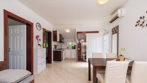 A modern kitchen with white cabinets, a dining area with a wooden table and white chairs, and a visible air conditioning unit. - House Ljerka (Barbat)