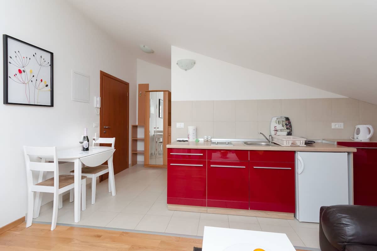 A modern kitchen with red cabinets, a white countertop, and a small refrigerator. There is a dining area with a white table and chairs.