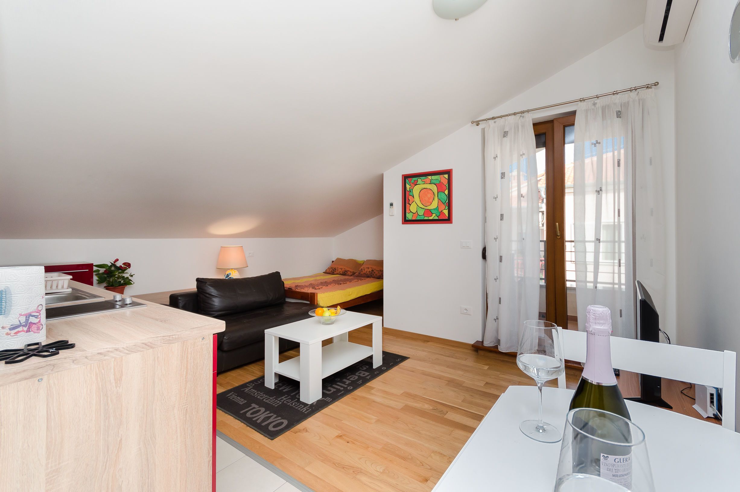 Uma sala de estar moderna com um sofá, uma mesinha branca e um espaço de refeições com uma cadeira e uma taça de vinho.