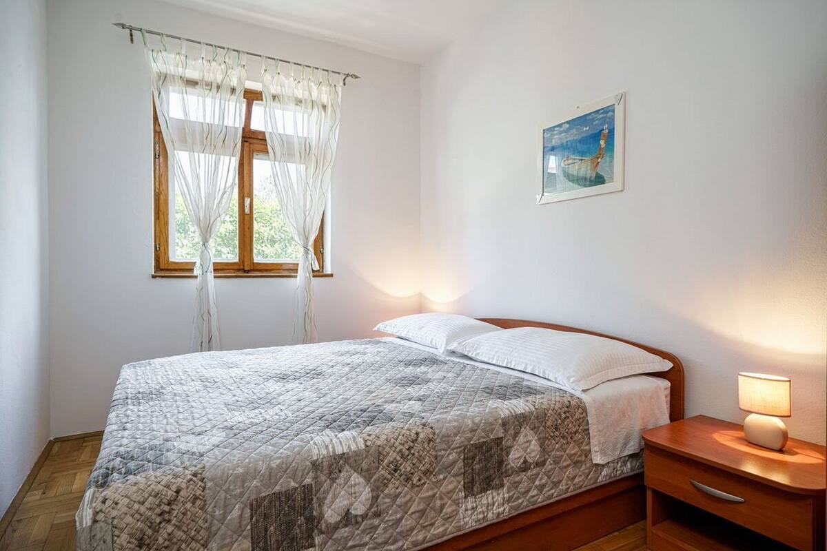 A bedroom with a bed, a nightstand, a window with sheer curtains, and a painting on the wall.