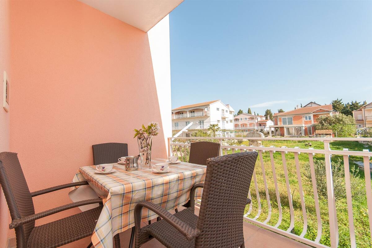 A balcony with a table set for two, wicker chairs, and a view of neighboring buildings.
