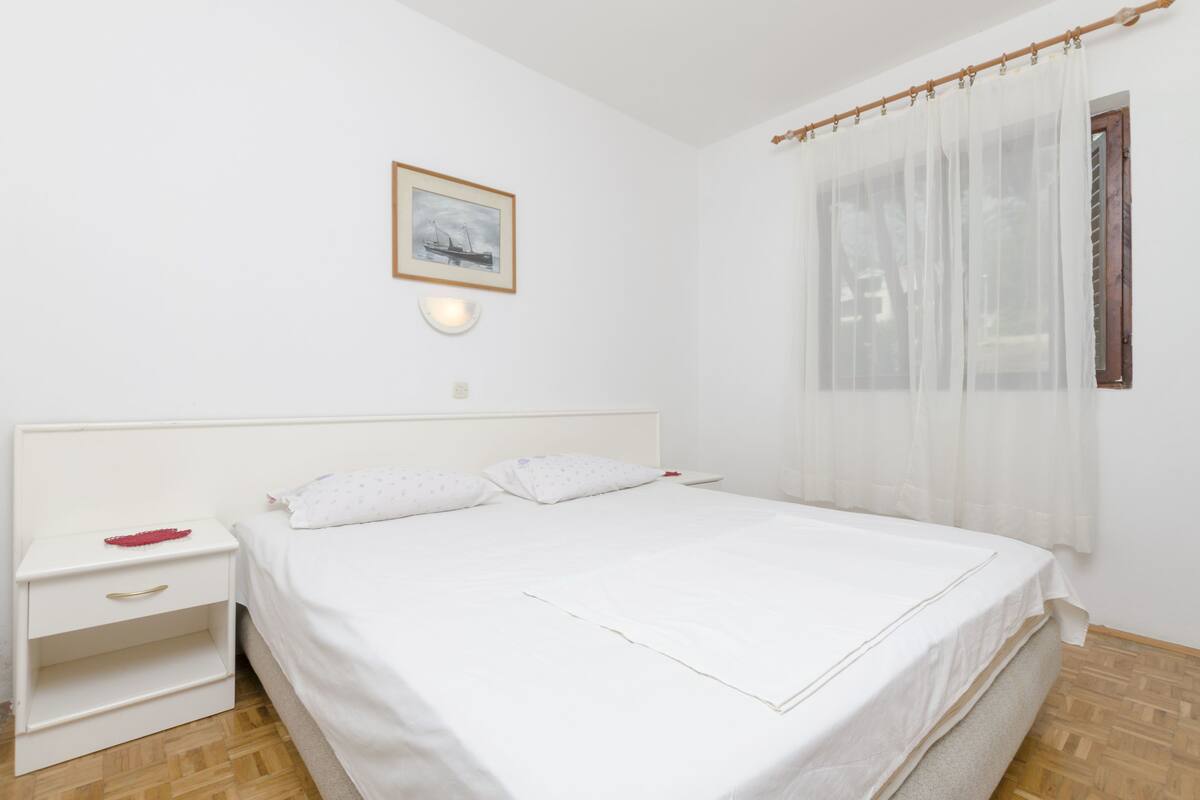 A neatly made bed with white bedding, a bedside table with a drawer, a wall-mounted light, and a window with sheer curtains.