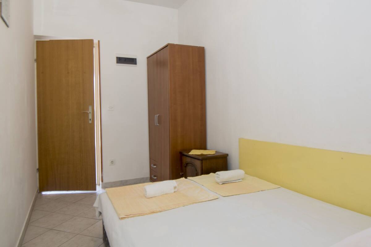 A hotel room with a bed, a wooden wardrobe, a small table, and a door leading to another room.
