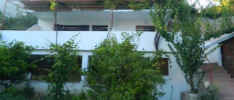 A two-story building with a stone wall in front, a staircase on the right, and greenery around.