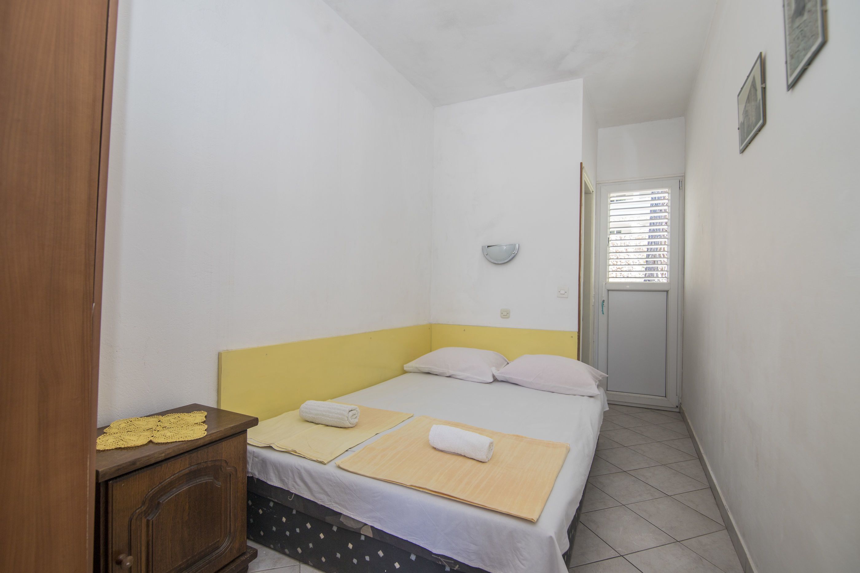 A small, narrow room with a bed, a wooden cabinet, and a window with blinds.