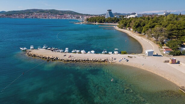 Eine Küstenlandschaft mit Booten, die an einem Sandstrand vertäut sind, einem klaren Gewässer und einer entfernten Stadtansicht.