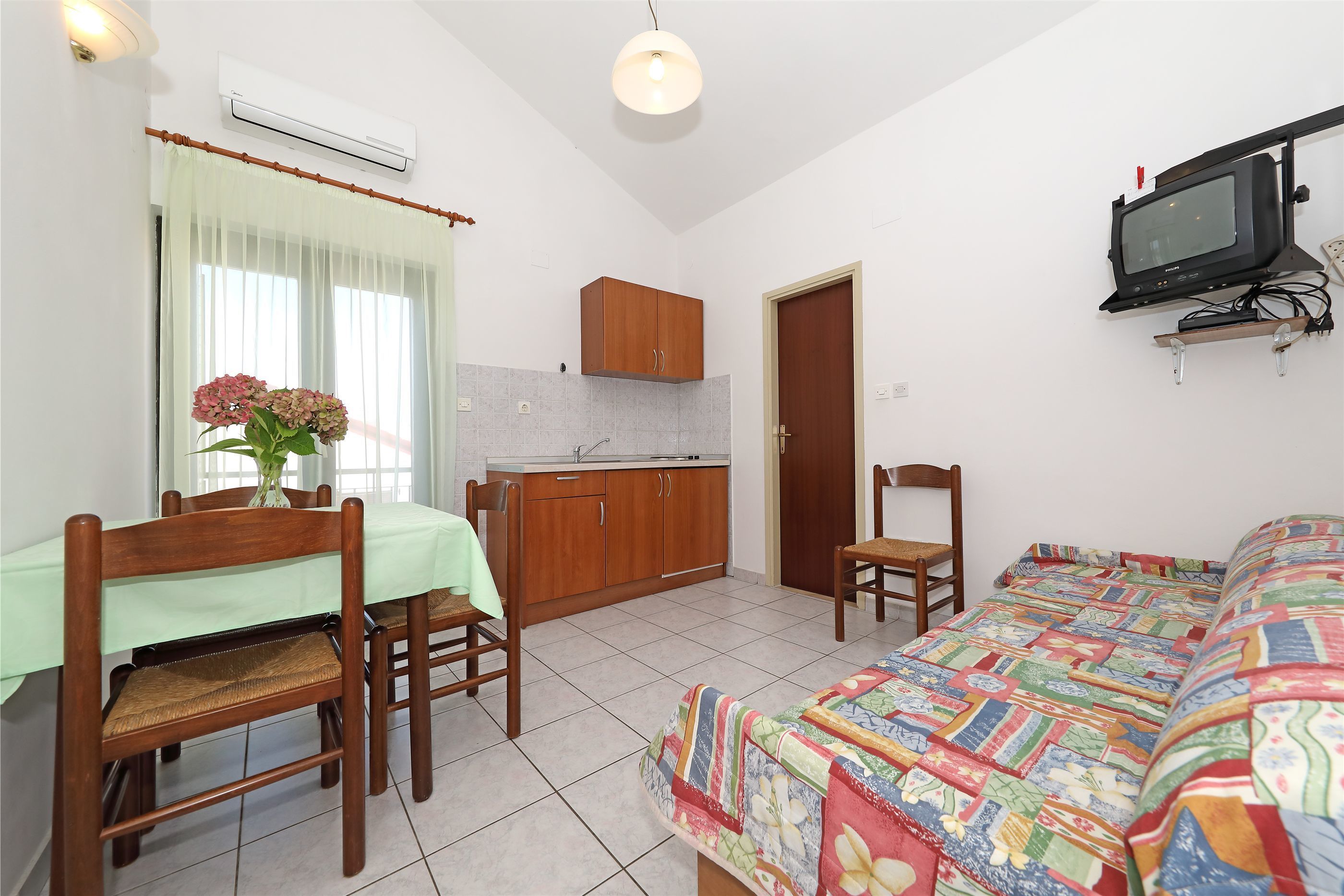 A compact room with a dining table, chairs, a bed, a small kitchenette, and a TV mounted on the wall.